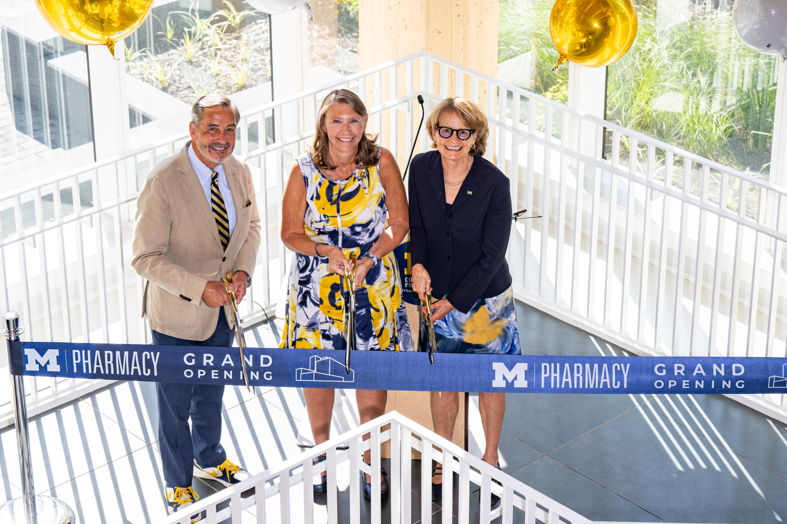 Three individuals participate in a ribbon-cutting ceremony for the M Pharmacy grand opening, standing on a staircase with balloons in the background.