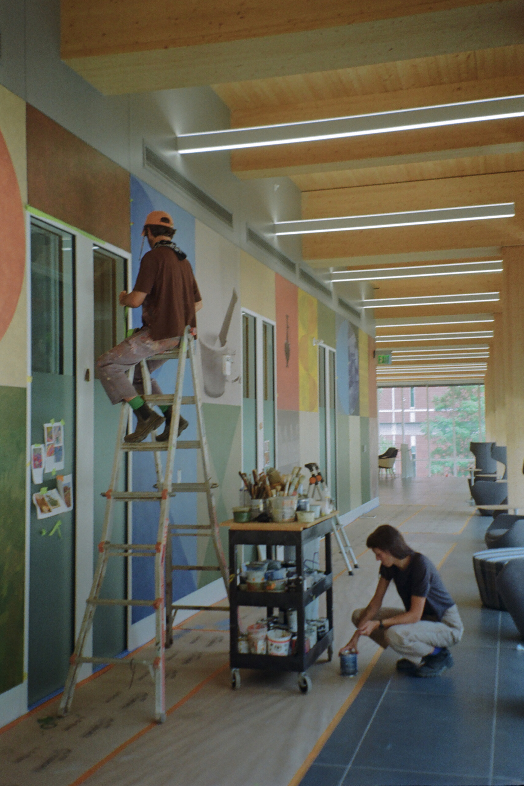 A person stands on a ladder painting a mural on a wall indoors, while another person kneels, working with paint supplies on the floor.
