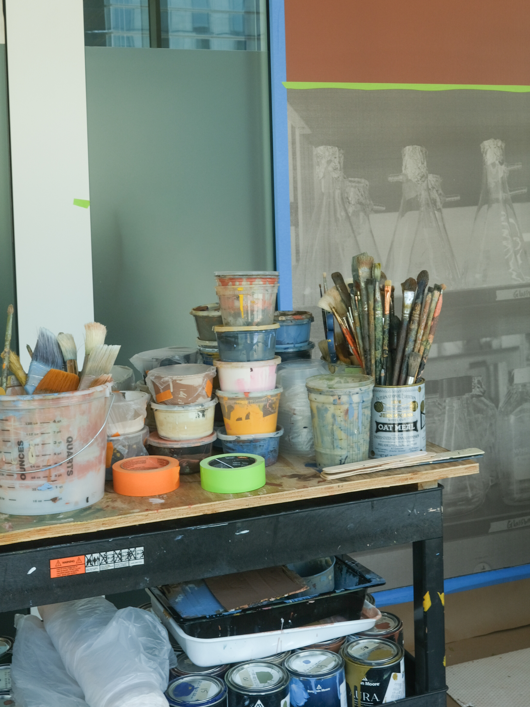 A workspace with various art supplies, including brushes, paint buckets, and rolls of tape, is arranged on a table. In the background, there's a large canvas featuring an unfinished mural.
