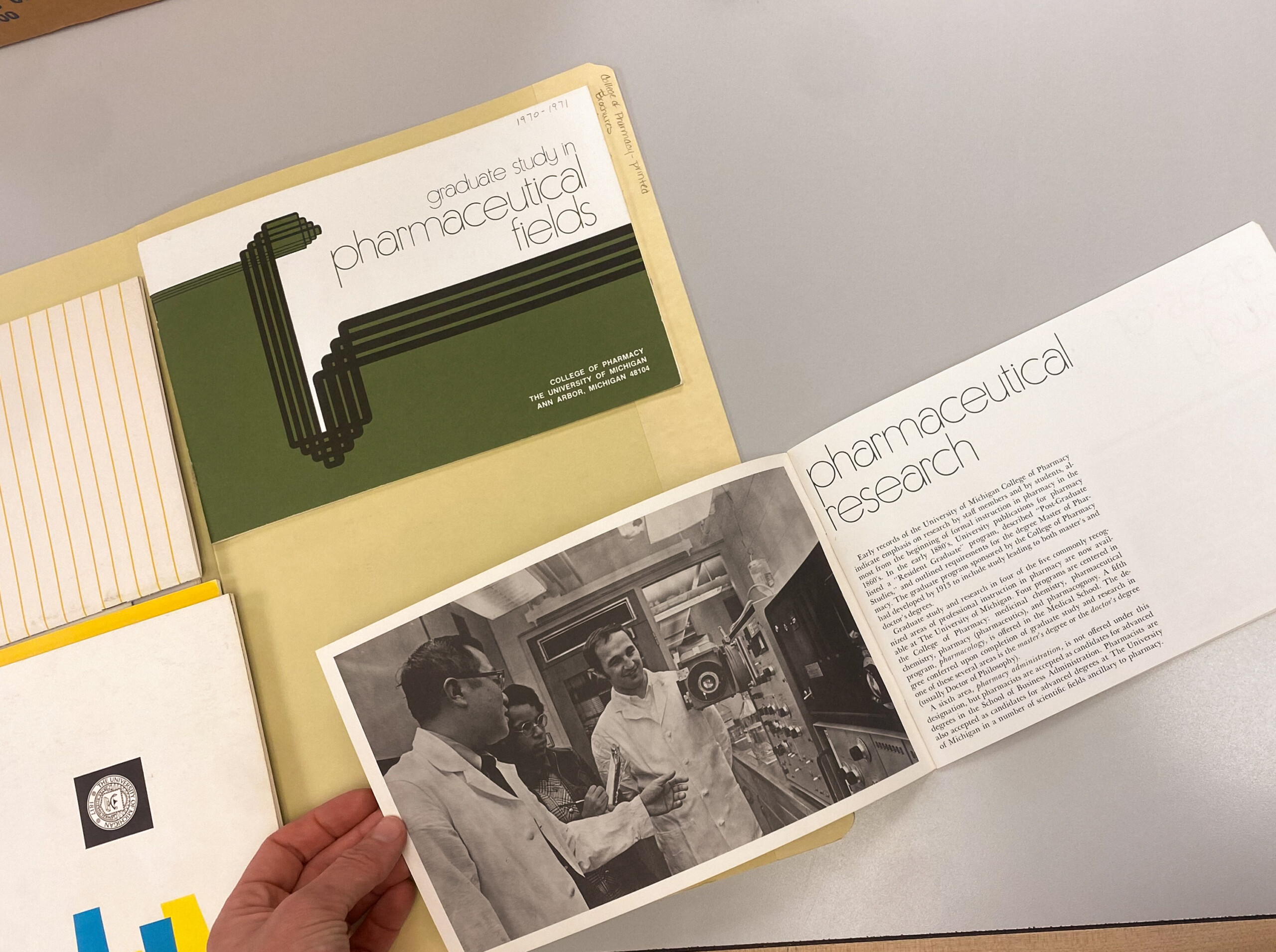 Folder labeled "Graduate Study in Pharmaceutical Fields." A person is holding a brochure titled "Pharmaceutical Research" featuring an image of three individuals in a laboratory setting.