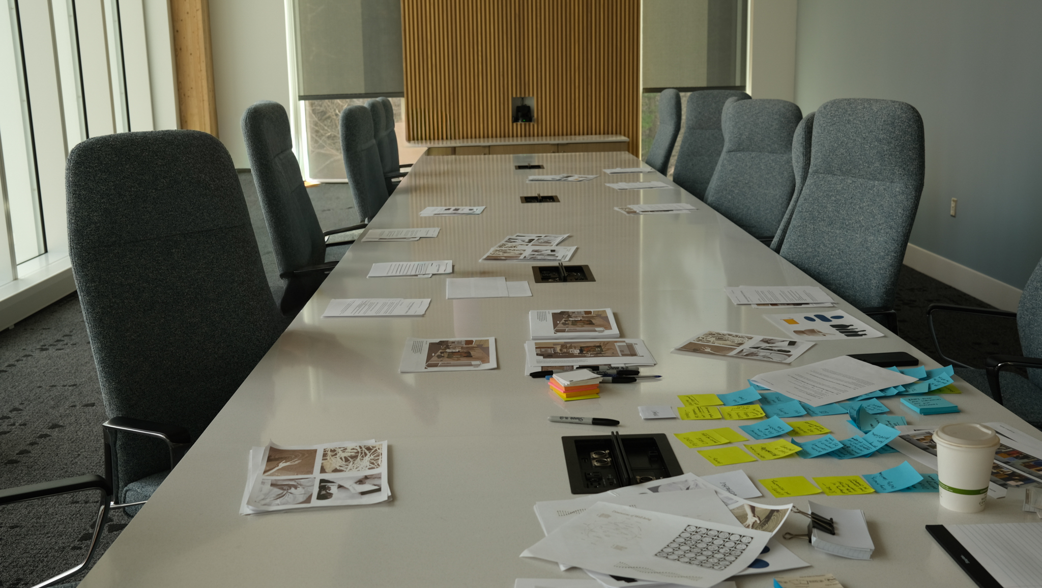A long conference table surrounded by office chairs, covered in various documents, photographs, sticky notes, and stationery.