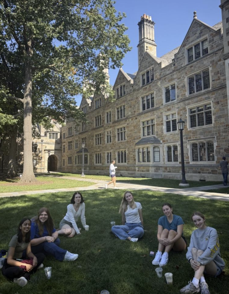 Group of women sitting on the grass in front of an academic building on University of Michigan Campus smiling