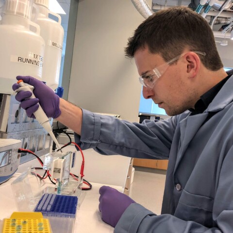 A person in a lab coat and safety goggles uses a pipette in a laboratory setting.